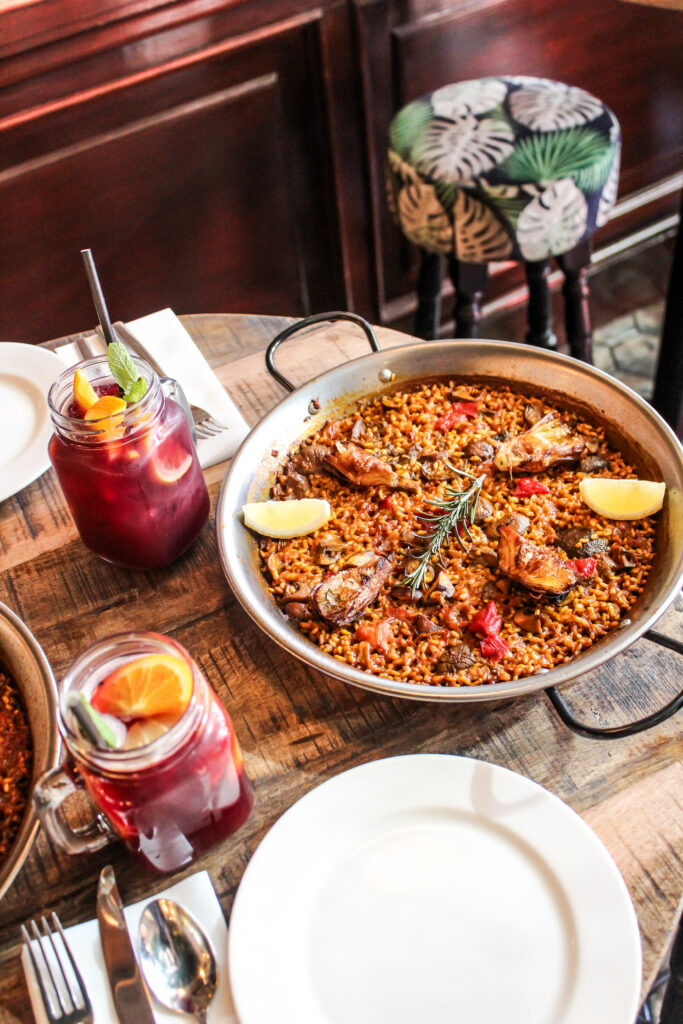 A pan of paella garnished with lemon wedges and rosemary sits on a wooden table, accompanied by two glasses of sangria with fruit slices and mint, empty plates, and cutlery.
