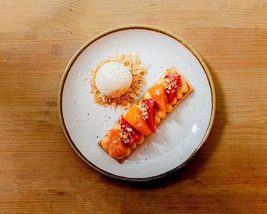 A rectangular fruit tart with colorful fruit slices and crumbles is plated beside a scoop of vanilla ice cream on a white dish, set on a wooden table.