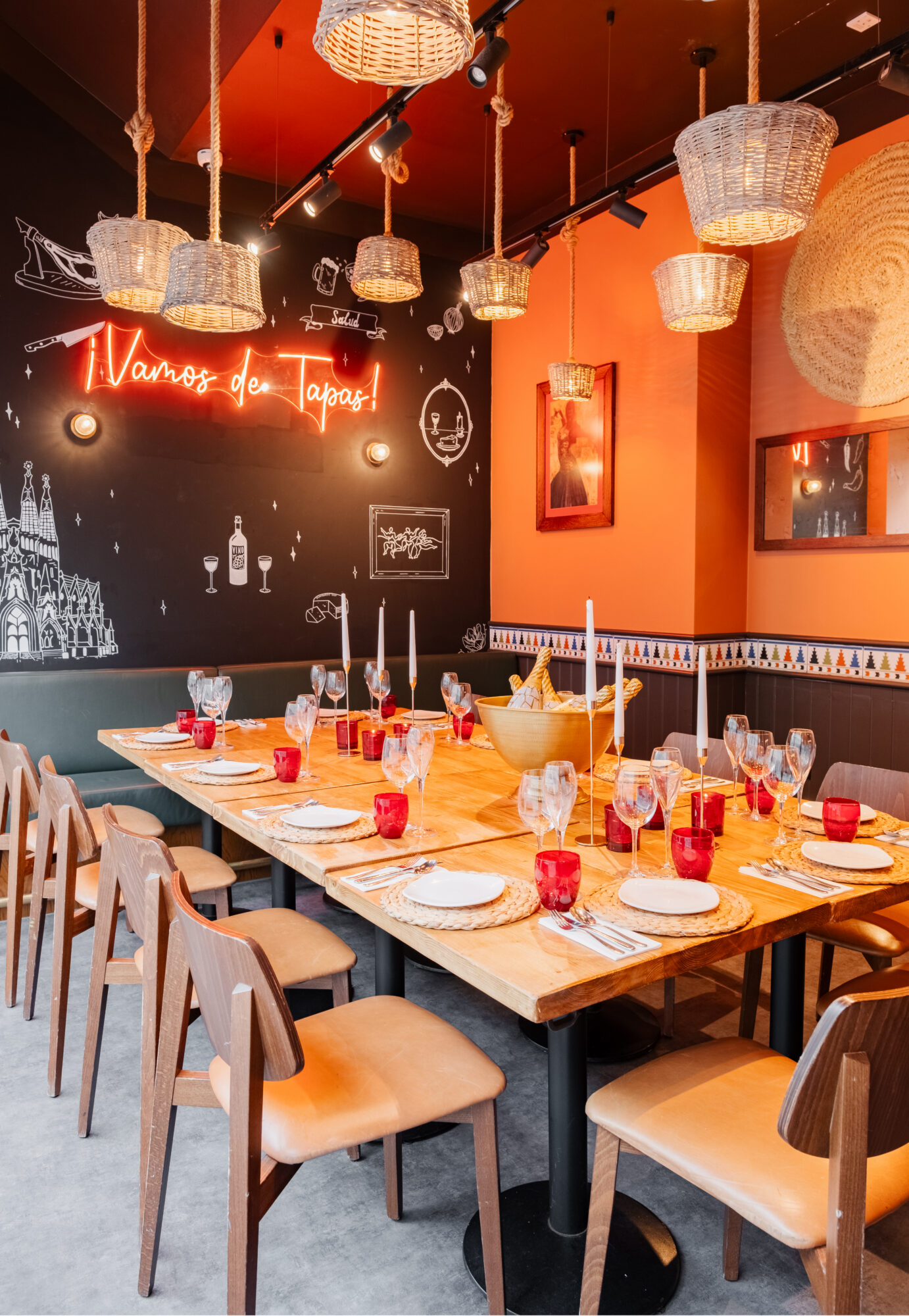 A restaurant dining area with a long wooden table set for a meal, featuring red glasses, plates, and candles. Warm lighting, woven lamps, and a black wall with Spanish-themed chalk art and a neon sign reading 