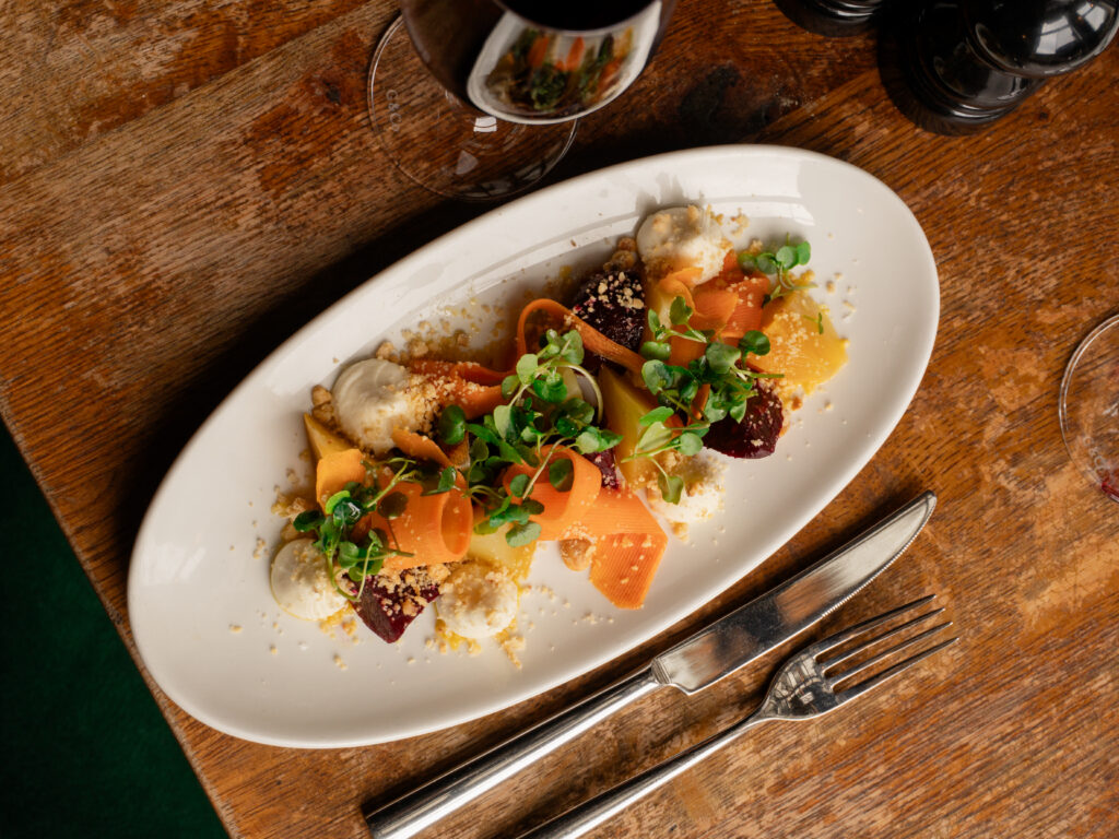 An overhead view of a plated gourmet salad with colorful vegetables, microgreens, crumbled cheese, and dollops of cream, served on a white oval dish with a knife and fork on a rustic wooden table. A glass of red wine is nearby.