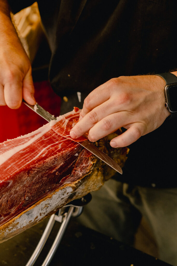 A person slices thin pieces of cured ham with a knife, holding the meat steady with one hand. The ham rests on a stand, and the person wears a black shirt and a smartwatch.