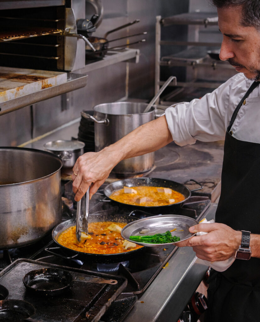 A chef in a commercial kitchen uses tongs to cook food in a pan on the stove while holding a plate of fresh herbs, with pots and other cookware visible in the background.