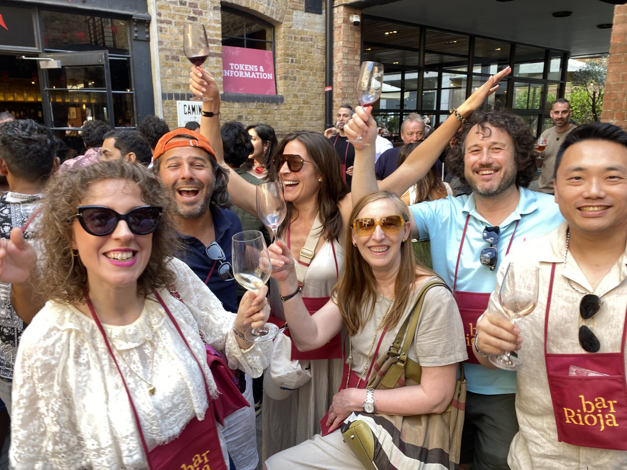 A group of six people smiling and raising wine glasses at an outdoor event. They wear maroon aprons with 
