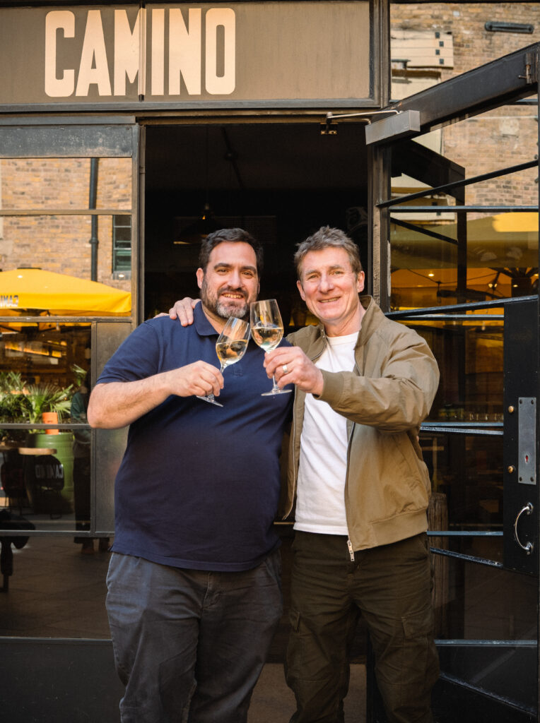 Two men smiling and clinking glasses of white wine while standing in front of a restaurant entrance with a sign that reads 