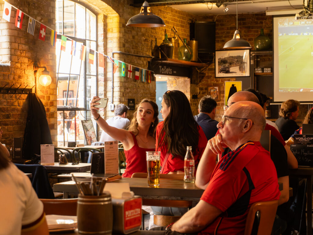 Two women take a selfie at a pub table with drinks, while other people watch a soccer match on a big screen. The pub is decorated with international flags and has brick walls and warm lighting.