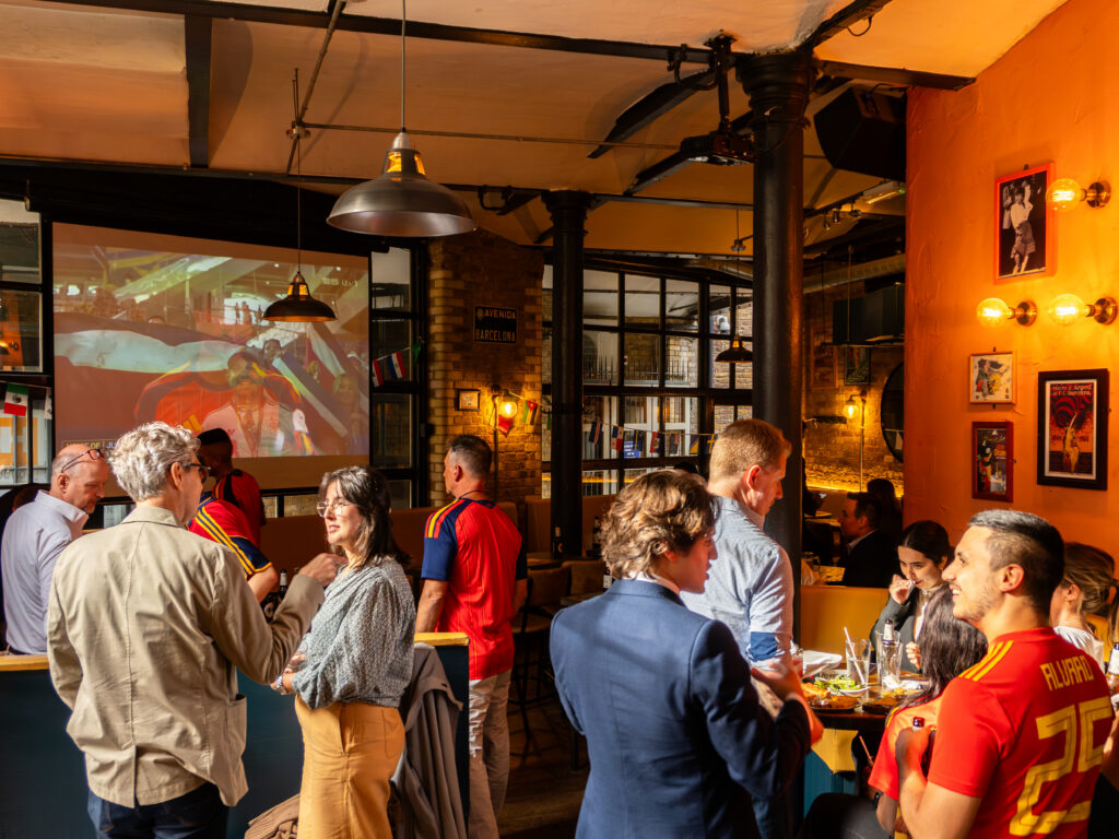 A group of people socialize in a warmly lit bar, some wearing red sports jerseys. A soccer match is projected on the wall. The atmosphere is lively and casual, with food and drinks on tables.