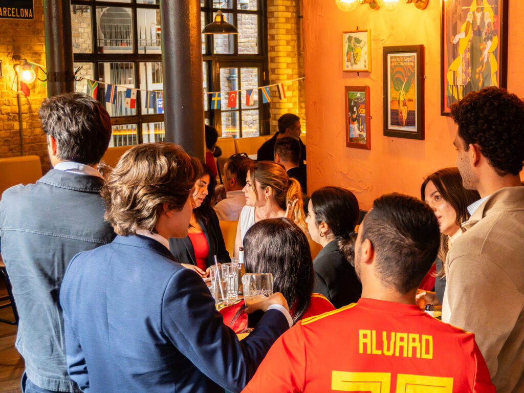 A group of people, some dressed in formal wear and one in a red soccer jersey, gather and socialize in a warmly lit bar or café decorated with framed pictures and banners.