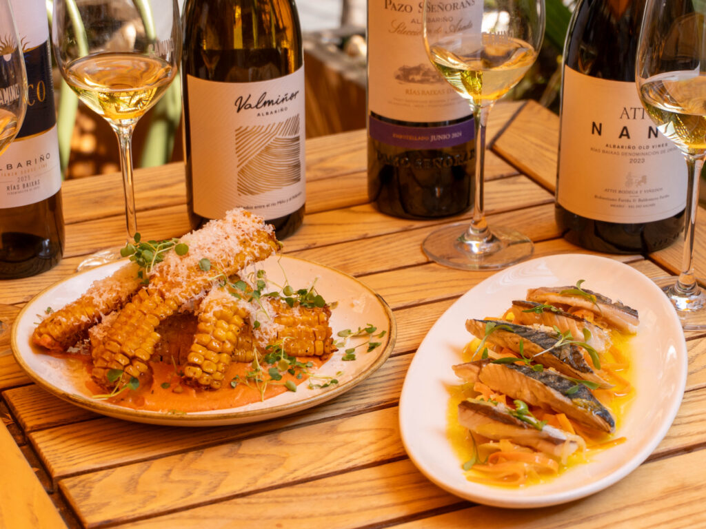 Two plates of food on a wooden table: grilled corn with grated cheese and herbs, and sliced fish with sauce, accompanied by wine bottles and glasses with white wine.