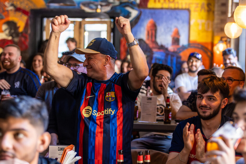 A man in a Barcelona soccer jersey cheers with arms raised in a crowded bar as others around him smile and clap, celebrating while watching a game.