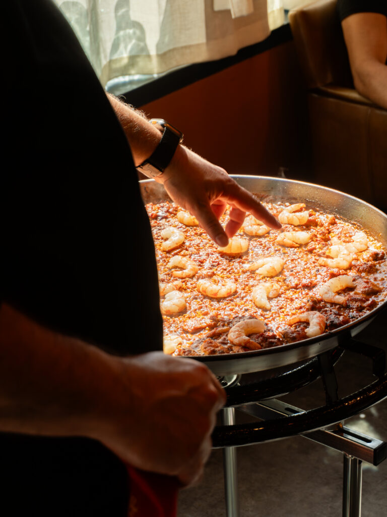 A person adds shrimp to a large pan of paella cooking on a stove, with sunlight coming through a window in the background.
