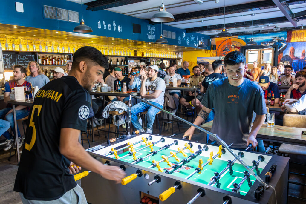 Two young men play foosball in a lively bar filled with people sitting at tables, enjoying drinks and watching the game. The bar has shelves of bottles, a colorful mural, and a vibrant, social atmosphere.