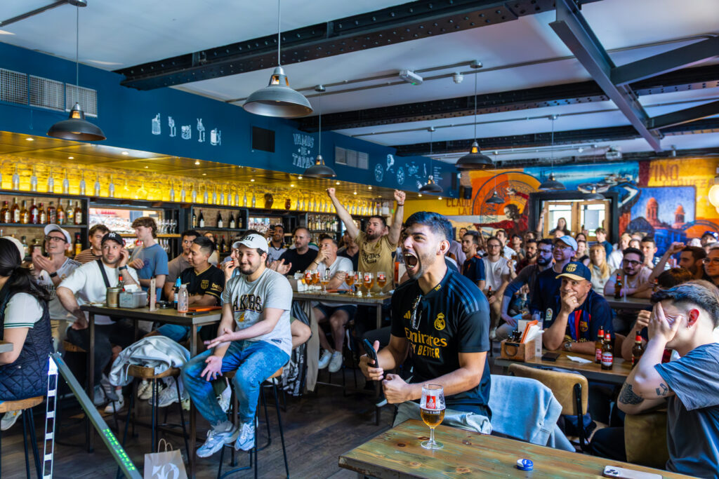 A lively crowd in a bar watches a sports event on TV; some people cheer and react excitedly while others sit and watch intently, drinks and snacks on the tables.