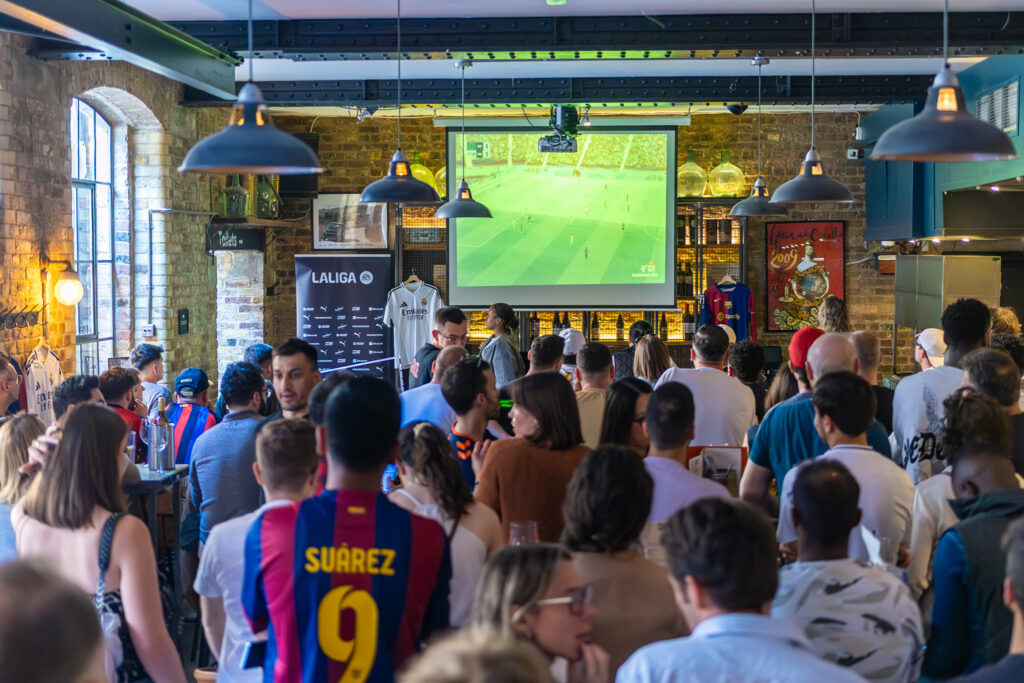 A crowd of people in a bar watch a soccer match projected on a screen. Many wear soccer jerseys, including one with “Suárez 9” in yellow. The atmosphere is lively, with fans standing and talking.