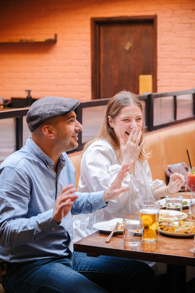 Two people sit at a restaurant table, laughing and enjoying drinks and food. The man wears a cap and blue shirt, while the woman in white covers her mouth as she laughs. The atmosphere is casual and cheerful.