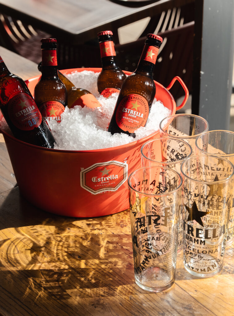 A red Estrella Damm bucket filled with ice and five beer bottles sits on a table next to four empty branded beer glasses in sunlight.