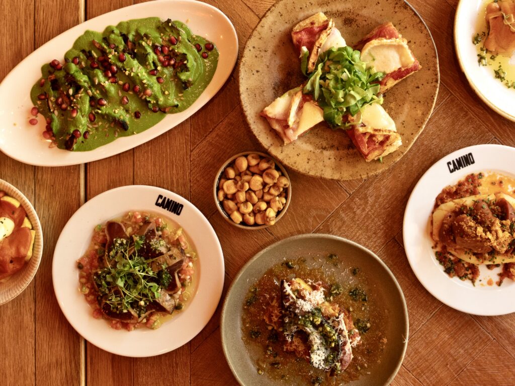 A wooden table set with assorted Spanish tapas, including green sauce-drizzled vegetables, grilled bread with greens, marinated nuts, and plated seafood and meat dishes.