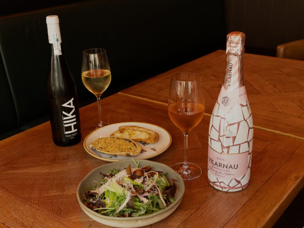 A table with salad, a slice of bread, a wine glass, and a bottle of white wine on one side, and sparkling wine with a pink-label bottle and a glass of rosé on the other.