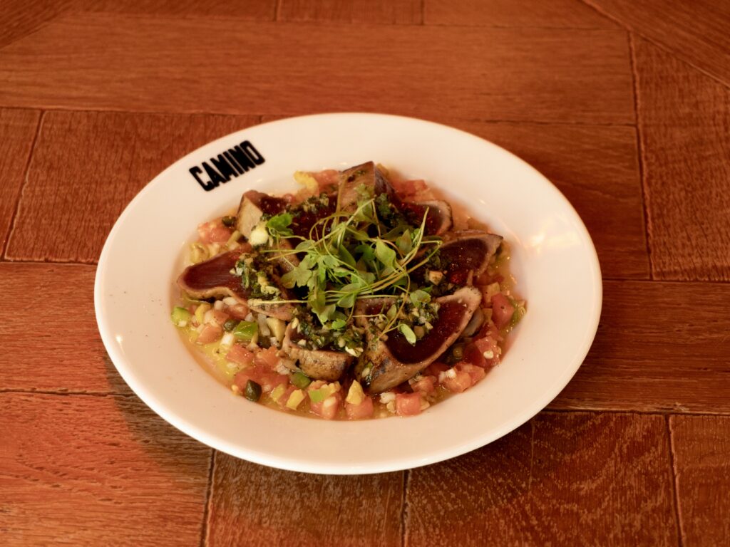 A white plate labeled “Camino” holds seared tuna slices on a bed of diced vegetables, topped with microgreens, placed on a wooden table.