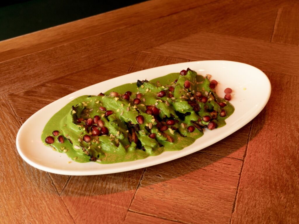 Oval white plate with sliced vegetables covered in green sauce, garnished with pomegranate seeds and herbs, placed on a wooden table.