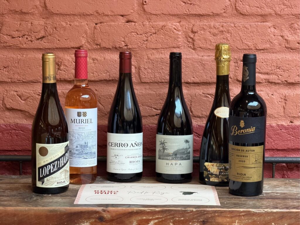 Six bottles of wine, including reds, rosé, and sparkling, are lined up on a wooden table against a red brick wall. A tasting mat with circles for glasses is placed in front of the bottles.