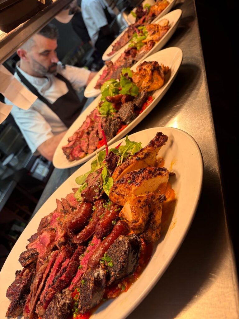 Three plates filled with assorted grilled meats, garnished with greens, are lined up on a metal counter. A chef in the background prepares food in a busy kitchen.